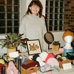 Indoor craft market stall displaying handmade knitted hats, scarves and accessories arranged in baskets and on tables. A person wearing glasses and a light-coloured jumper stands behind the stall. Mannequin heads model knitted hats, while potted plants, a mirror and a sign reading “Suzy Makes Stuff” decorate the display. The stall is set against glass-block windows and brick walls, creating a warm community market atmosphere.