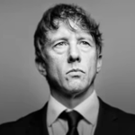 Black-and-white studio portrait of a person in a dark suit jacket, white shirt and dark tie against a plain grey background. The person is shown from the chest up, facing forward with their gaze directed slightly upward. Soft lighting creates contrast across the face and clothing, while the background fades smoothly from darker grey at the edges to lighter grey near the centre.