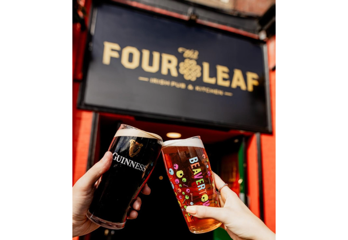 A pint of beer and a pint of Guinness being clinked together under The Four Leaf Irish Bar & Kitchen's sign. 