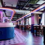 Modern bar interior with a sleek, curved counter in the foreground, lit by bright red, blue, and white neon strip lights tracing the ceiling. Behind the bar are drink fridges and shelves. The space features high tables with upholstered stools, patterned wall panels, and geometric lighting fixtures. A section of ceiling greenery adds contrast to the dark metal framework. Large windows on the right flood the room with daylight, highlighting polished wooden floors and a decorative tiled area near the bar.