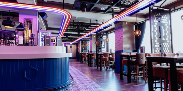 Modern bar interior with a sleek, curved counter in the foreground, lit by bright red, blue, and white neon strip lights tracing the ceiling. Behind the bar are drink fridges and shelves. The space features high tables with upholstered stools, patterned wall panels, and geometric lighting fixtures. A section of ceiling greenery adds contrast to the dark metal framework. Large windows on the right flood the room with daylight, highlighting polished wooden floors and a decorative tiled area near the bar.