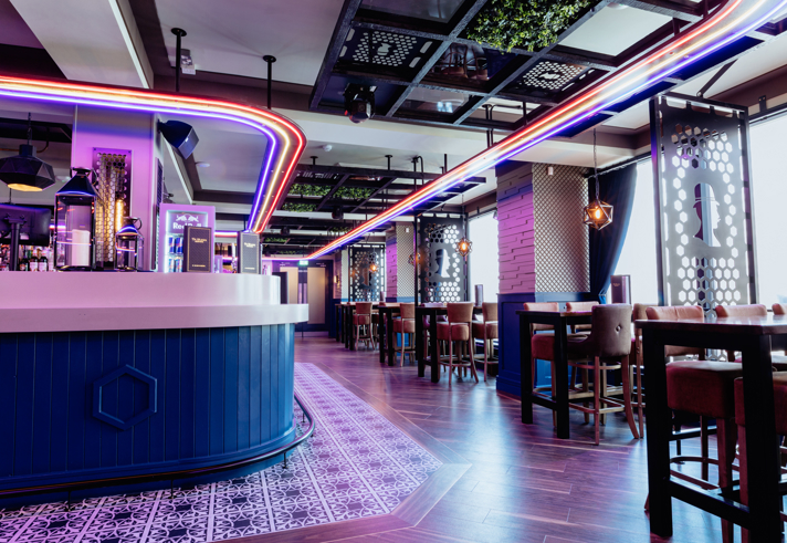 Modern bar interior with a sleek, curved counter in the foreground, lit by bright red, blue, and white neon strip lights tracing the ceiling. Behind the bar are drink fridges and shelves. The space features high tables with upholstered stools, patterned wall panels, and geometric lighting fixtures. A section of ceiling greenery adds contrast to the dark metal framework. Large windows on the right flood the room with daylight, highlighting polished wooden floors and a decorative tiled area near the bar.