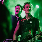 Two musicians perform on stage under green lighting. One is playing a black electric bass guitar and wearing a sleeveless shirt with patches, while the other stands slightly behind holding a microphone. The background features stage equipment and a smoky atmosphere, creating a vibrant live concert setting.