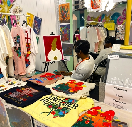 Pop-up market stall featuring vibrant printed T-shirts, framed artwork, and a painting on an easel. The table displays multiple colorful designs, and additional art pieces are hung on the wall behind.