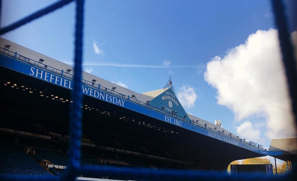 View of Hillsborough Stadium, home of Sheffield Wednesday FC, taken from behind a goal net. The blue seating and main stand are visible, with signage reading ‘Sheffield Wednesday’ and ‘Est. 1867’ under a bright blue sky with scattered clouds.