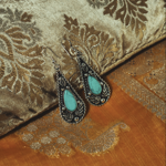 Pair of ornate teardrop earrings with turquoise stones set in intricate silver metalwork, displayed on luxurious gold and bronze patterned fabric.