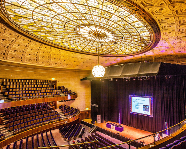 The Oval Hall at Sheffield City Hall. Grand auditorium with tiered seating and a large stage featuring a projection screen. The ornate ceiling has an intricate geometric design with a central stained-glass dome and a hanging light fixture. The stage is framed by black curtains, and the seating area includes multiple balconies with curved railings.