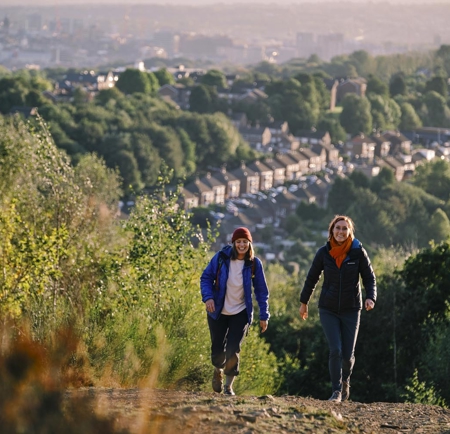 Two ramblers are walking up a hill. Down in the valley below them you can see rows of terrace houses and on the horizon you can see Sheffield city centre.