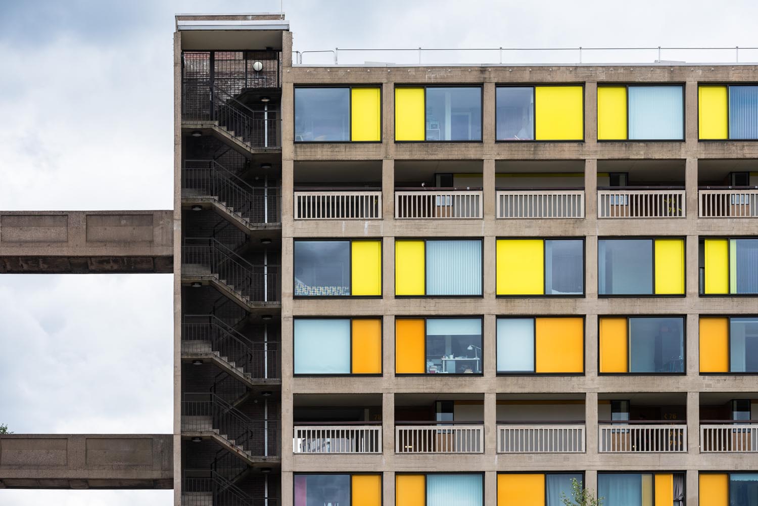 A section of a modernist apartment building with a concrete exterior and a grid of square windows in bright yellow, orange, and light blue panels. The left side features an open stairwell with metal railings and a connecting walkway. The overall design is geometric and colourful, contrasting with the cloudy sky in the background.