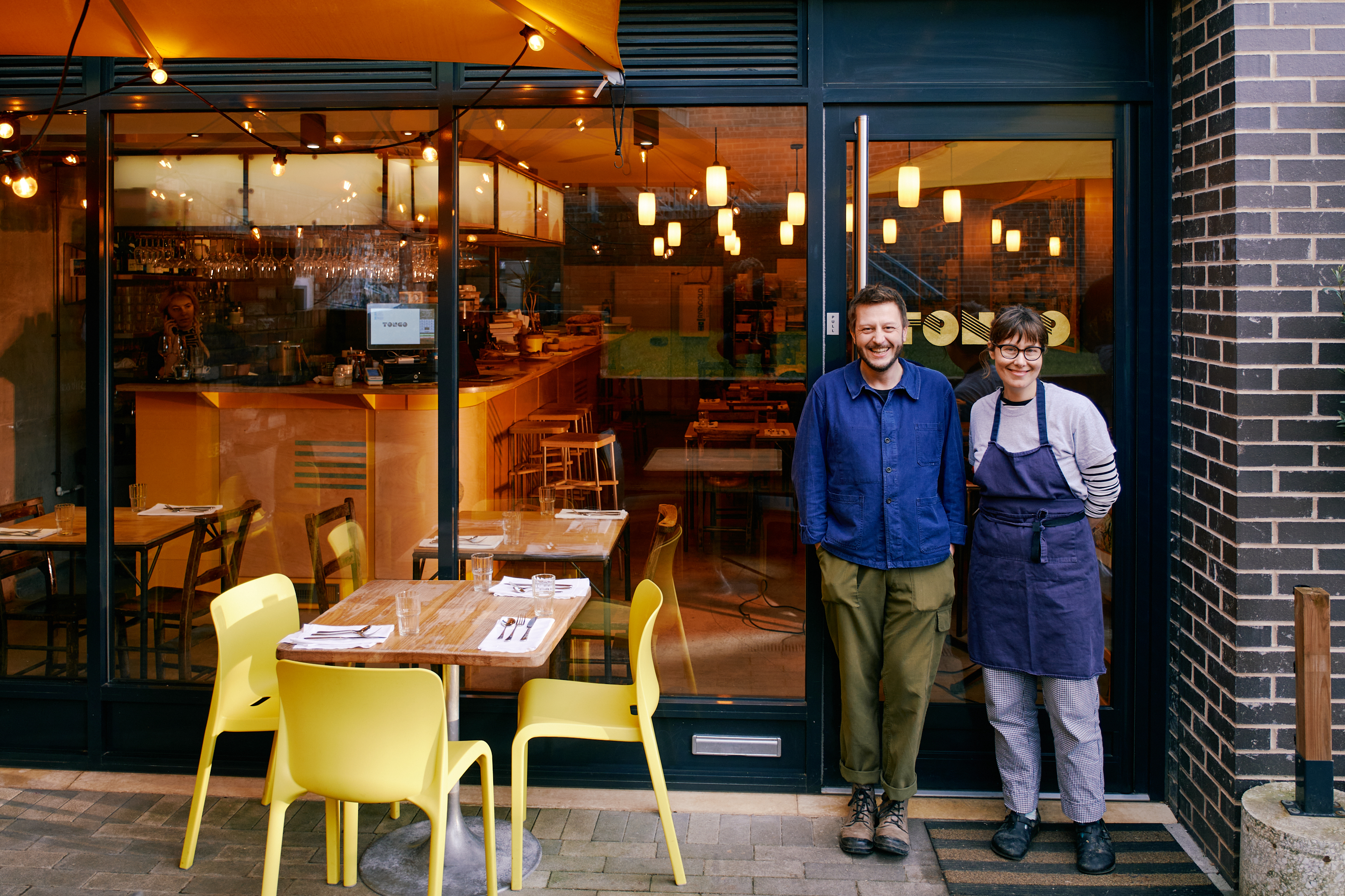 A smiling man and woman stand outside their restaurant, smiling at the camera.