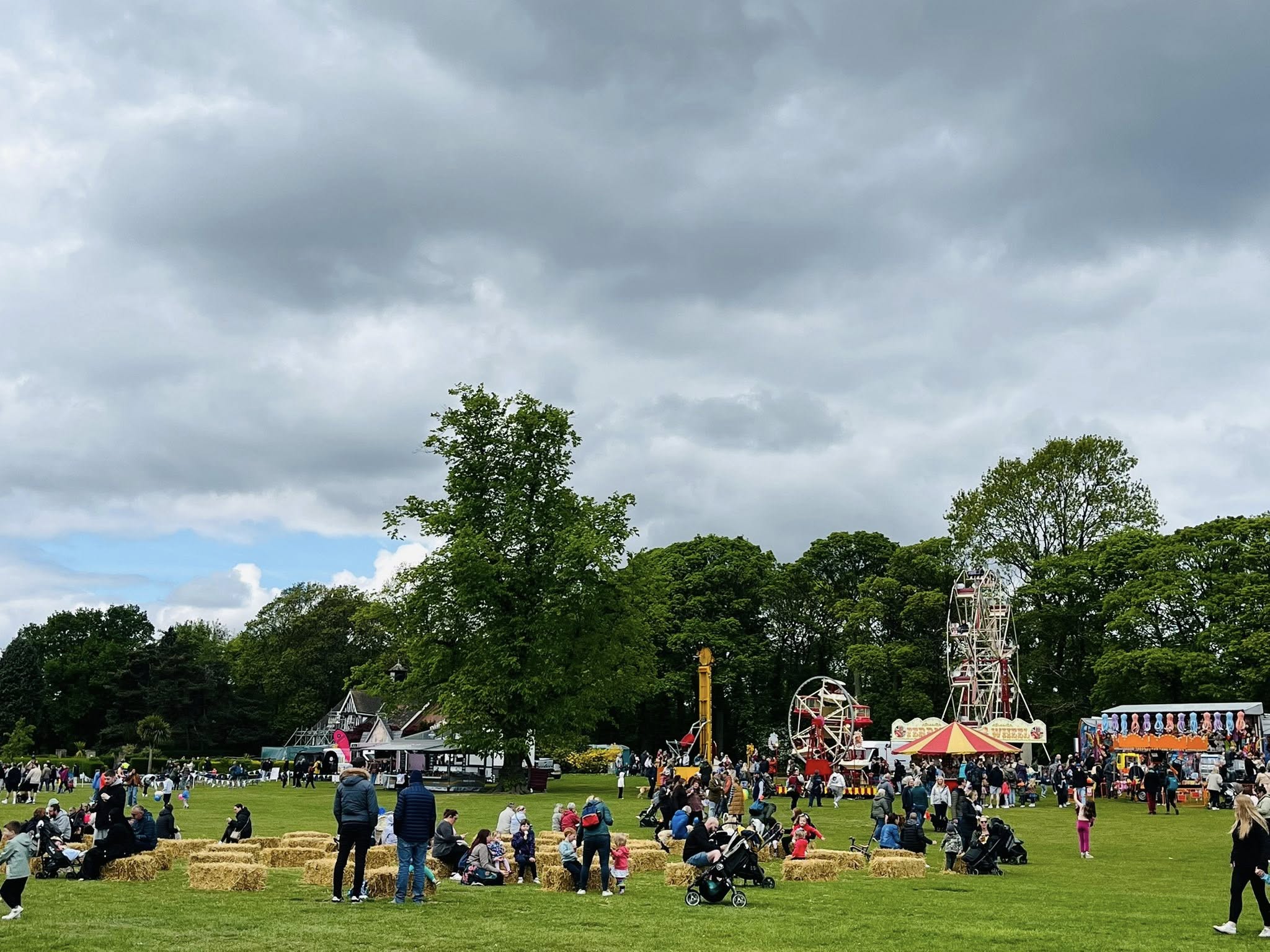 A busy outdoor fair on a large grassy field with groups of people sitting on hay bales and walking around. In the background are fairground rides, including two Ferris wheels and a tall yellow ride, along with colourful stalls and a striped tent. Trees line the edge of the field under a cloudy sky, creating a lively yet overcast atmosphere.