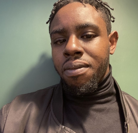 Person wearing a dark turtleneck sweater and a black jacket, standing against a plain green wall. A small round fixture is visible on the wall near the top right of the image.