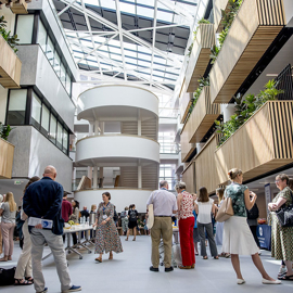 Spacious modern atrium with a glass ceiling and natural light, featuring multiple floors with wooden slatted balconies and green plants. A group of people is gathered in the open central area, some standing near tables with refreshments. The design includes curved white staircases and sleek architectural details, creating a bright and contemporary setting.