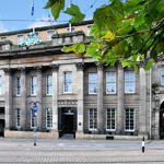 The exterior of Cutlers' Hall. A historic stone building with neoclassical architecture featuring tall columns, large rectangular windows, and ornate detailing above the entrance. The facade includes decorative carvings and a crest at the top center. Tram tracks run along the street in front, and green leaves from a nearby tree frame the upper part of the image.