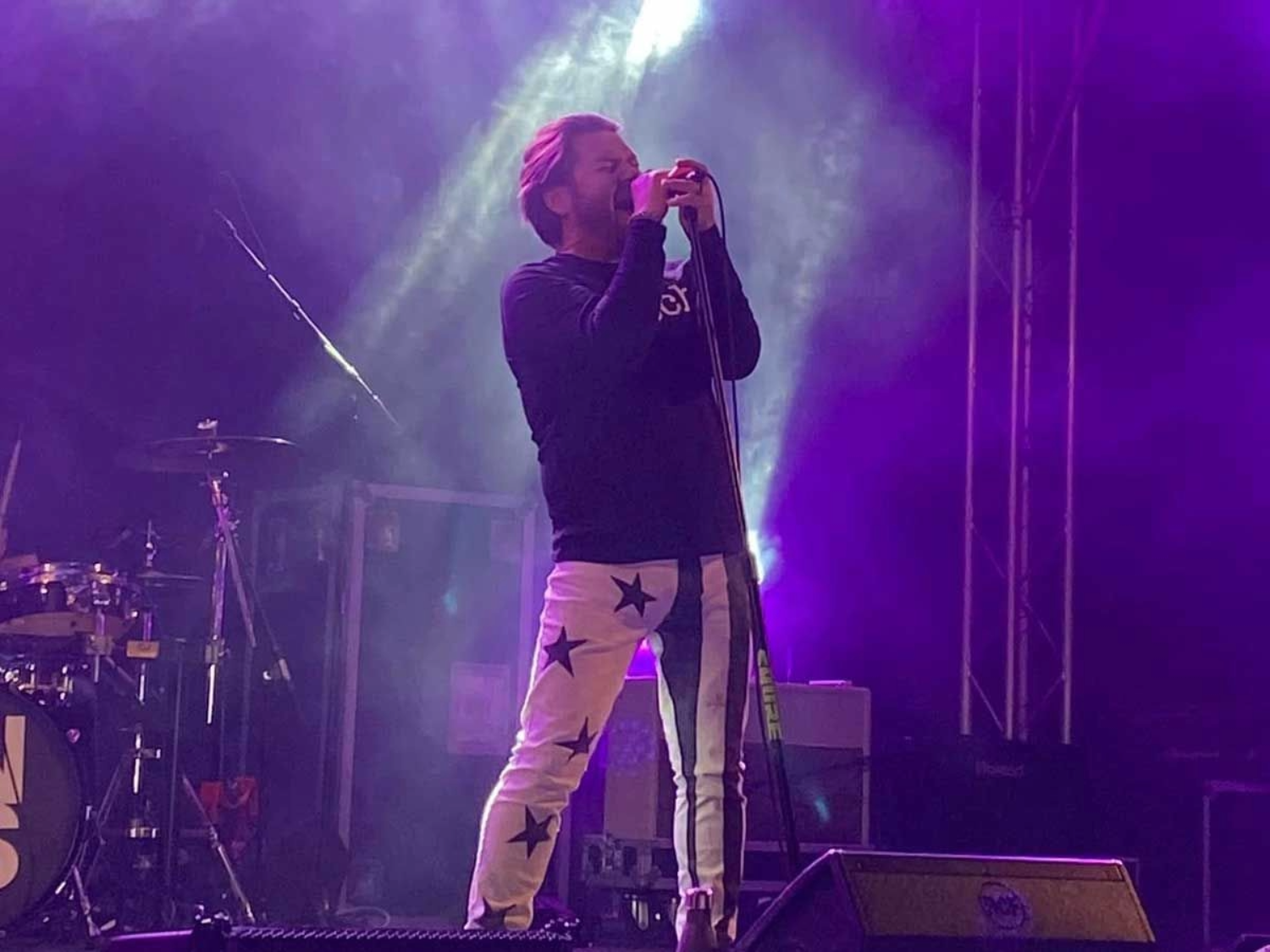A performer stands on stage singing into a microphone, wearing a black long-sleeve top and white pants decorated with large black stars. Bright purple and white stage lights create a dramatic backdrop, with drums and equipment visible in the background.