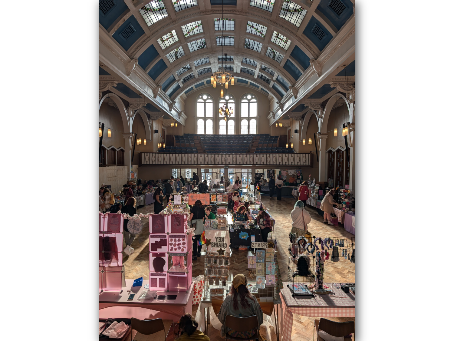 Indoor craft market in a grand hall with a high arched ceiling, large windows, and hanging lights. Several stalls with colourful displays of prints, jewellery, and handmade items line the wooden floor, with people browsing and sitting behind tables. Pink shelving units and signs like “Prints” and “Keychains” are visible in the foreground. Balcony seating and ornate architectural details frame the space.