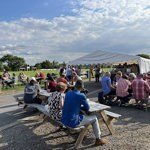 A sunny outdoor scene at a beer festival with groups of people sitting at wooden picnic benches on a paved area beside a large grassy field. A white marquee tent is set up in the background with people gathered around it. The sky is bright with scattered clouds.