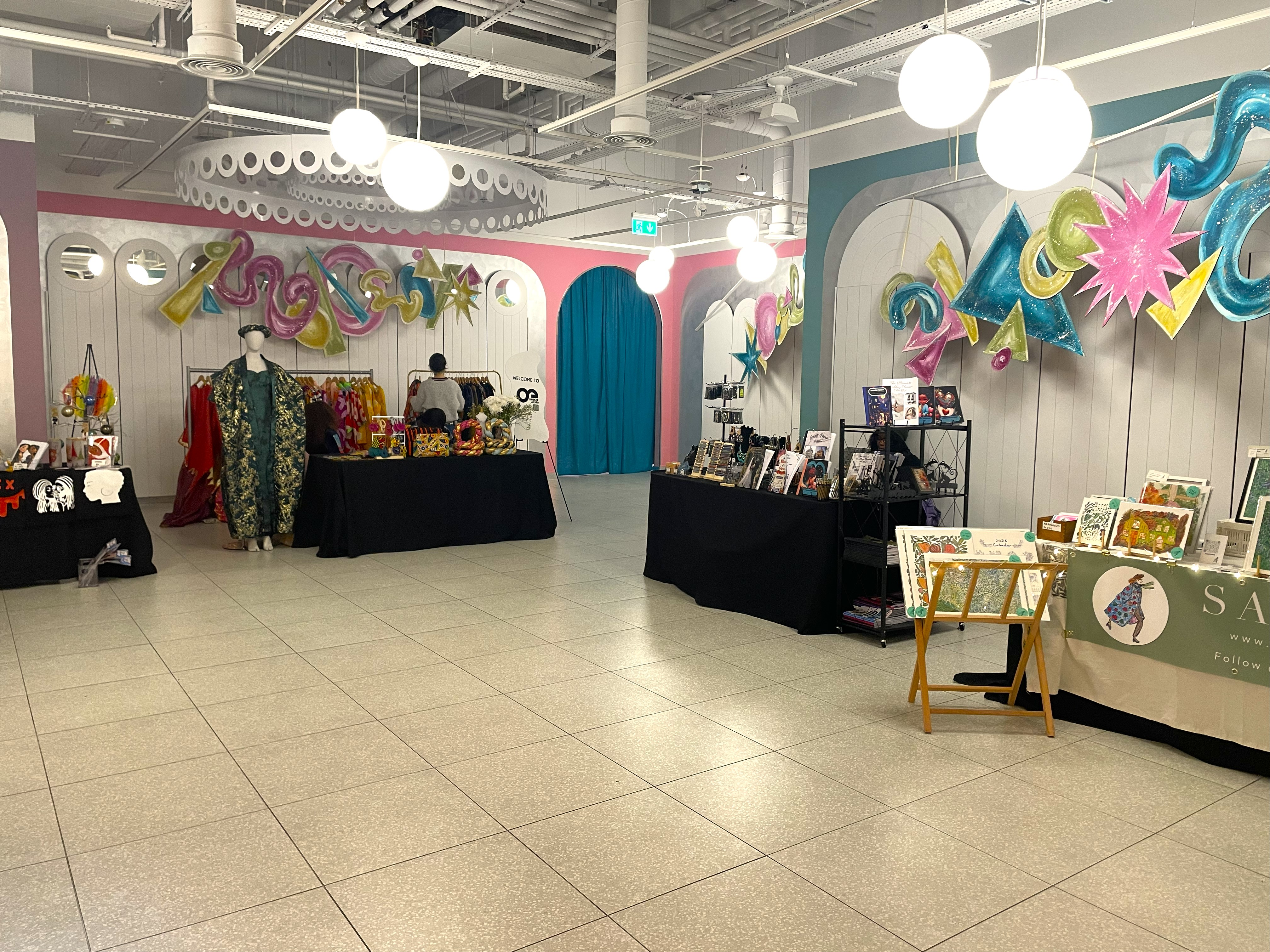 Wide view of a pop-up market interior with tables covered in black cloth displaying handmade crafts, artwork, and clothing. The space features vibrant wall decorations in pink, teal, and yellow with large hanging lights.
