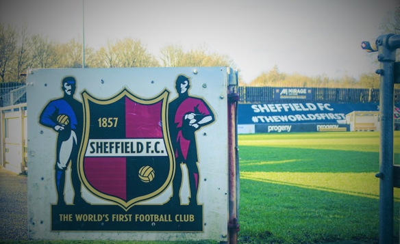 Signboard featuring the Sheffield F.C. crest with text ‘1857 Sheffield F.C.’ and ‘The World’s First Football Club,’ flanked by illustrations of two footballers. In the background, a football pitch and advertising boards display ‘Sheffield FC #TheWorldsFirst.’ Trees and sky are visible beyond the stands.