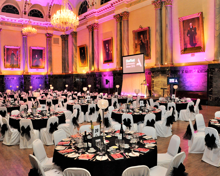 Elegant banquet hall with high ornate ceilings, chandeliers, and large portraits on the walls. Round tables are set for a formal dinner with white chair covers tied with black bows, black tablecloths, and red menus. A projector screen at the front displays “Sheffield Ambassador Dinner 2015.” Warm lighting with pink and orange uplights creates a vibrant atmosphere.