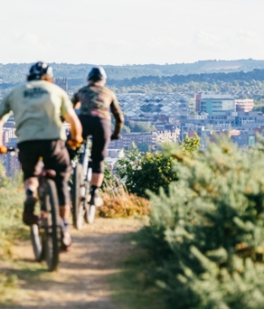 Two cyclists riding mountain bikes along a narrow dirt trail surrounded by green shrubs, overlooking a cityscape with modern buildings, cranes, and distant hills under a bright sky.