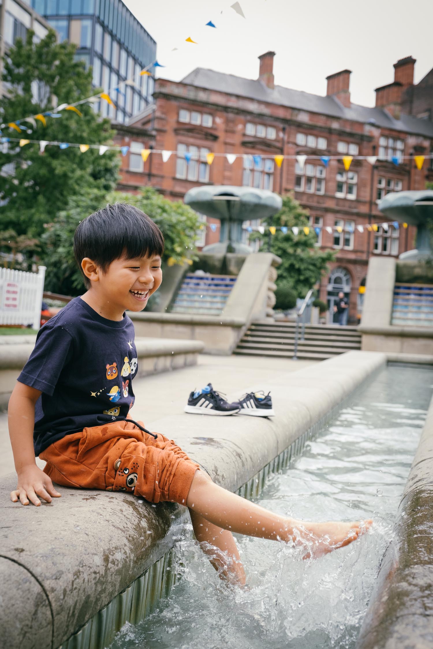 A child sits on the edge of a shallow water feature, splashing feet in the water. Shoes are placed on the stone ledge nearby. In the background, there are fountains, trees, and a historic red-brick building decorated with colourful bunting.