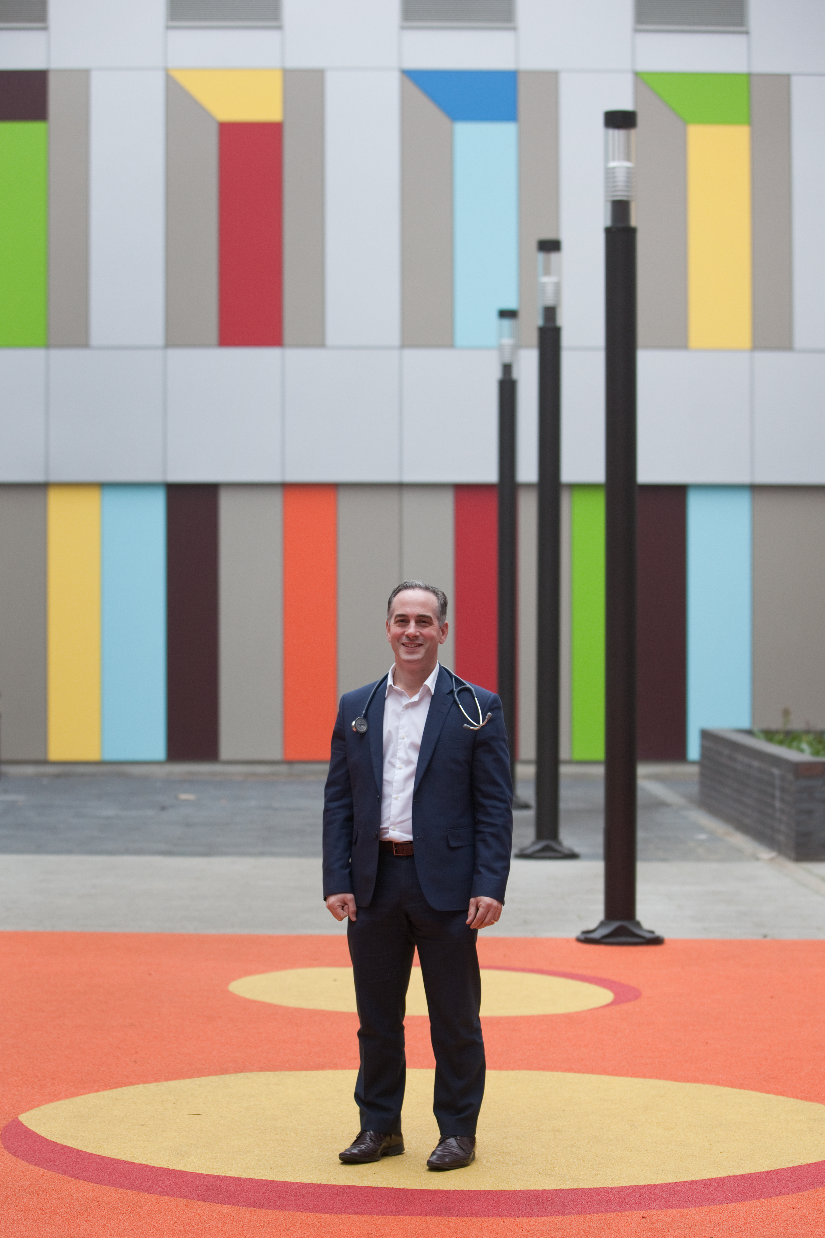 Paul Dimitri standing in a courtyard at the Sheffield Childrens Hospital. Person standing on an orange and yellow patterned outdoor surface in front of a modern building with a colorful vertical panel design in shades of green, blue, red, yellow, and gray. Several tall black lamp posts line the walkway, and the setting appears contemporary and urban.