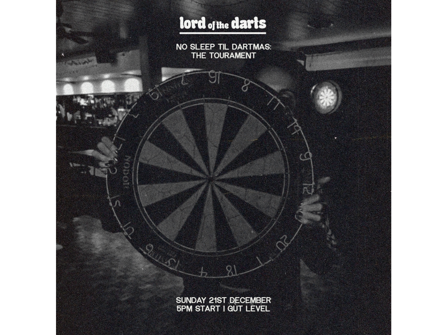Black-and-white promotional image for a darts event. A person holds a large dartboard in the centre, partially obscuring their face. The background shows a dimly lit bar with another dartboard on the wall. White text at the top reads “lord of the darts” followed by “NO SLEEP TIL DARTMAS: THE TOURNAMENT.” At the bottom, text states “SUNDAY 21ST DECEMBER, 5PM START | GUT LEVEL.” The overall design has a gritty, high-contrast aesthetic.