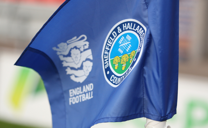 Close-up of a blue corner flag featuring the Sheffield & Hallamshire County FA crest with a white rose and crossed tools, alongside the England Football logo. The flag is positioned on a football pitch with blurred background.