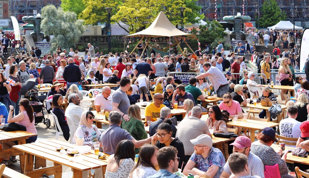 A huge crowd of people sit and stand around in a public square surrounded by street food stands on a sunny day 