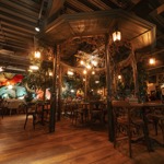 A warmly lit restaurant interior with wooden floors, wooden tables and chairs, large potted plants, and soft ambient lighting, creating a cosy, natural atmosphere.