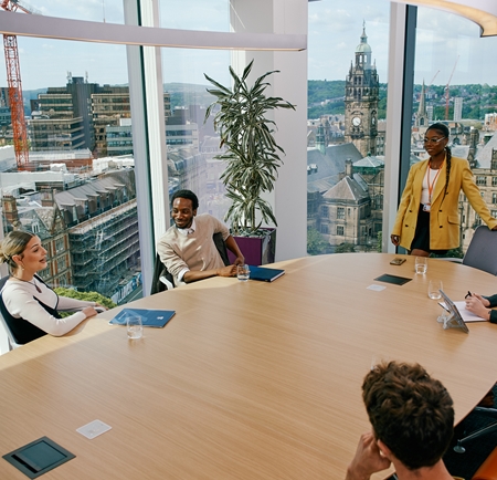 Four people, sat around a table in an office, are having a meeting. Through the pane glass windows Sheffield city centre is visible.