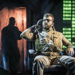  A dramatic stage scene features a person wearing a tactical-style beige outfit and eye patch, seated in a chair under theatrical lighting. A rope is draped across one shoulder. Behind, a shadowy figure stands in a doorway illuminated by orange light. To the right, a glowing digital-style backdrop displays vertical streams of green binary code. The set combines industrial and cyber-themed elements, creating a tense, futuristic atmosphere.