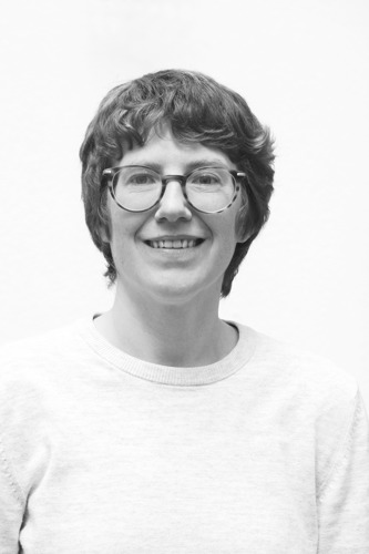 Black-and-white head-and-shoulders portrait of a person wearing round glasses and a light-coloured jumper against a plain background. The person faces the camera directly, with short wavy hair and a slight smile. The simple composition, even lighting and uncluttered backdrop create a clean, professional profile photograph.