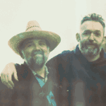 A picture of two men, one with his arm around the other, smiling.