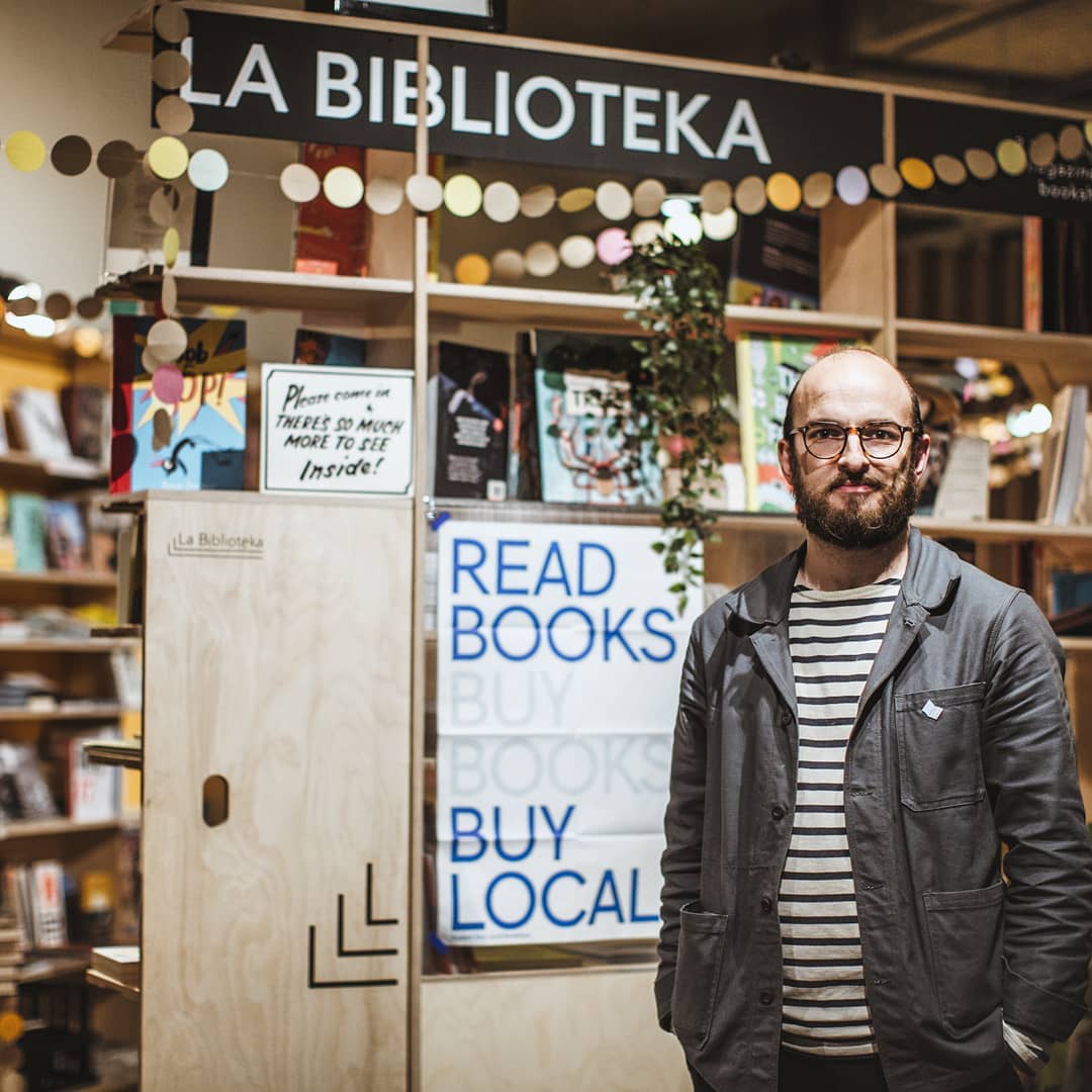 A man stands in a book shop, smiling at the camera.