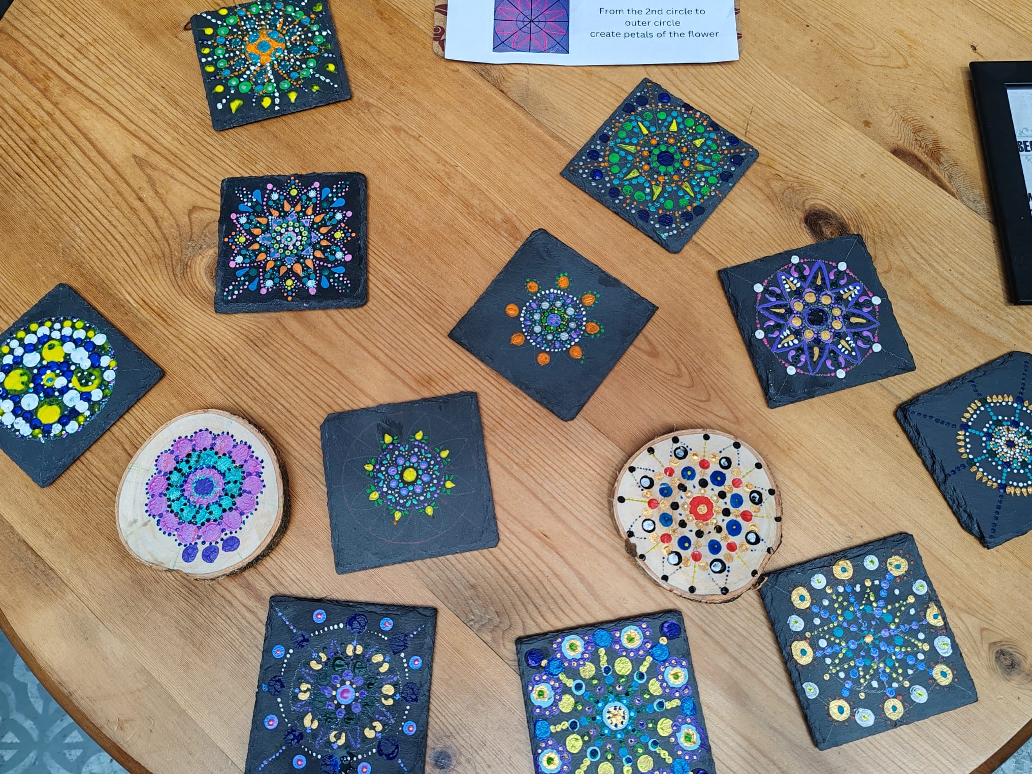 A wooden table displaying several hand-painted mandala-style coasters arranged in a loose circle. The designs feature colourful dotted patterns in blues, greens, yellows, purples and reds on dark square bases, with one round coaster decorated in multicoloured dots. A small instruction sheet is partially visible at the top.