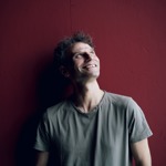 A person, smiling and looking up, stands against a dark red wall.