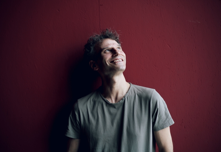 A person, smiling and looking up, stands against a dark red wall.