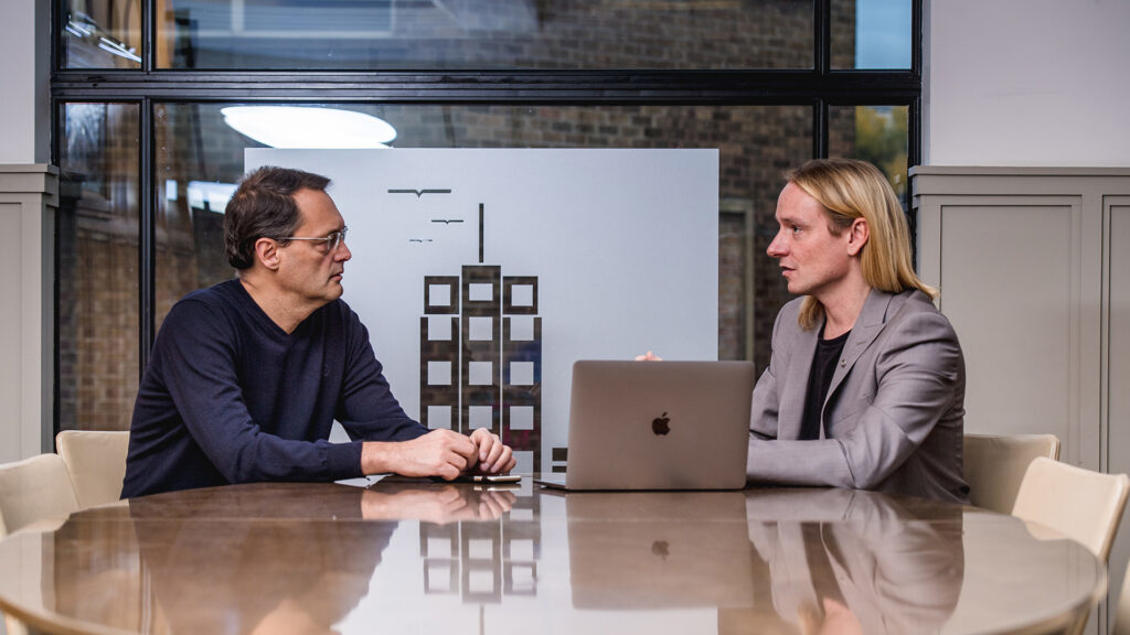Two men, smartly dressed, sat at a table talking. There is a laptop computer on the table.