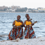 Two performers stand on a sandy shoreline beside the water, wearing matching bright yellow tops, colourful patterned skirts and headwraps. Each holds a traditional stringed musical instrument while performing outdoors. Waves roll in behind them, with a distant city skyline visible across the water. The image combines vibrant clothing with a coastal setting, creating a striking performance scene.