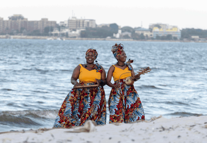 Two performers stand on a sandy shoreline beside the water, wearing matching bright yellow tops, colourful patterned skirts and headwraps. Each holds a traditional stringed musical instrument while performing outdoors. Waves roll in behind them, with a distant city skyline visible across the water. The image combines vibrant clothing with a coastal setting, creating a striking performance scene.