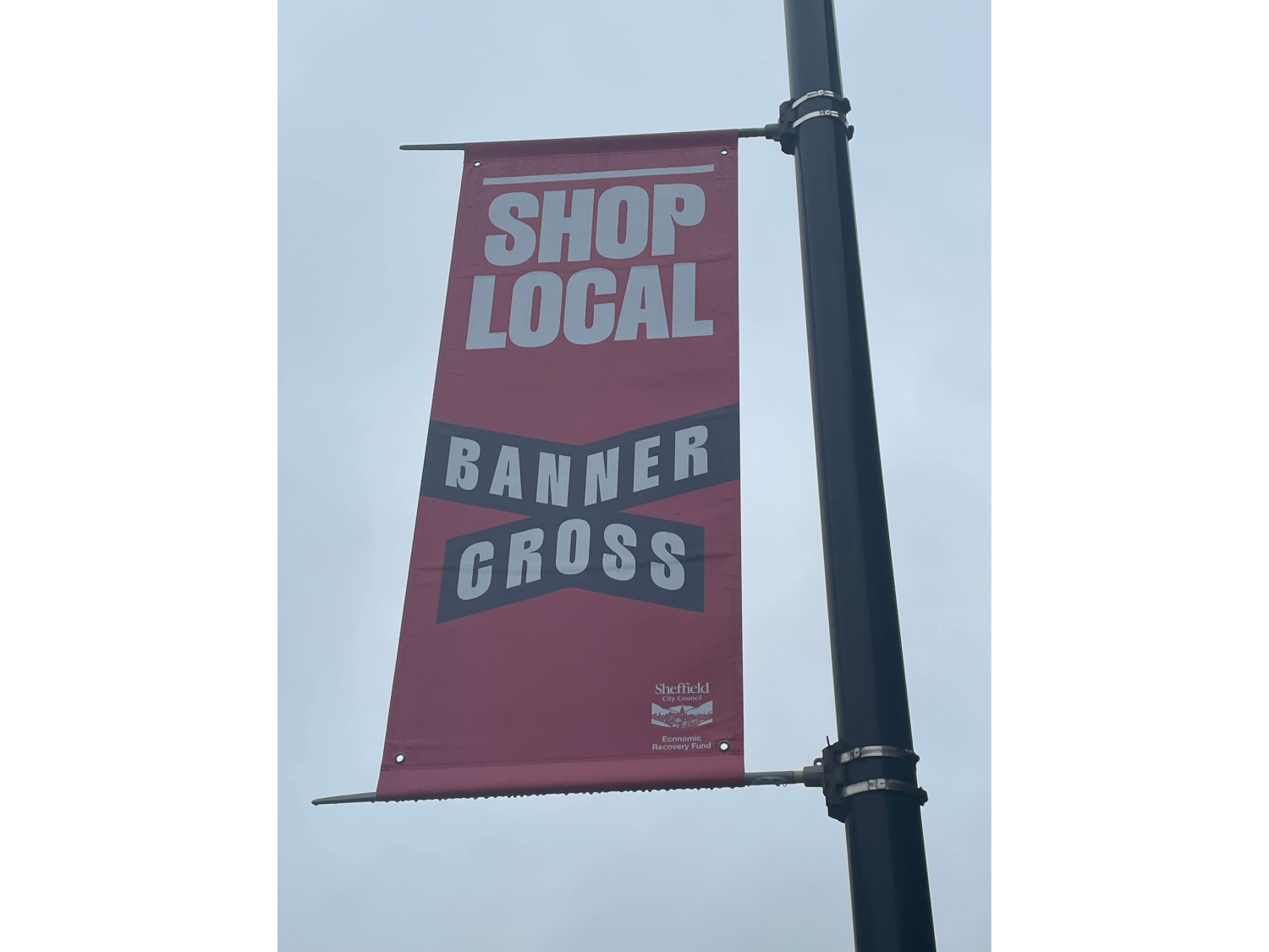 Vertical street banner attached to a black pole with the text ‘SHOP LOCAL’ in large letters and ‘BANNER CROSS’ in a diagonal design. The banner is red with white and black text, and includes a small Sheffield City Council logo at the bottom.
