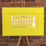 Contemporary artwork displayed on an easel, featuring a bright yellow frame and a central metallic panel with cut-out fork-like shapes, set against a brick wall background.