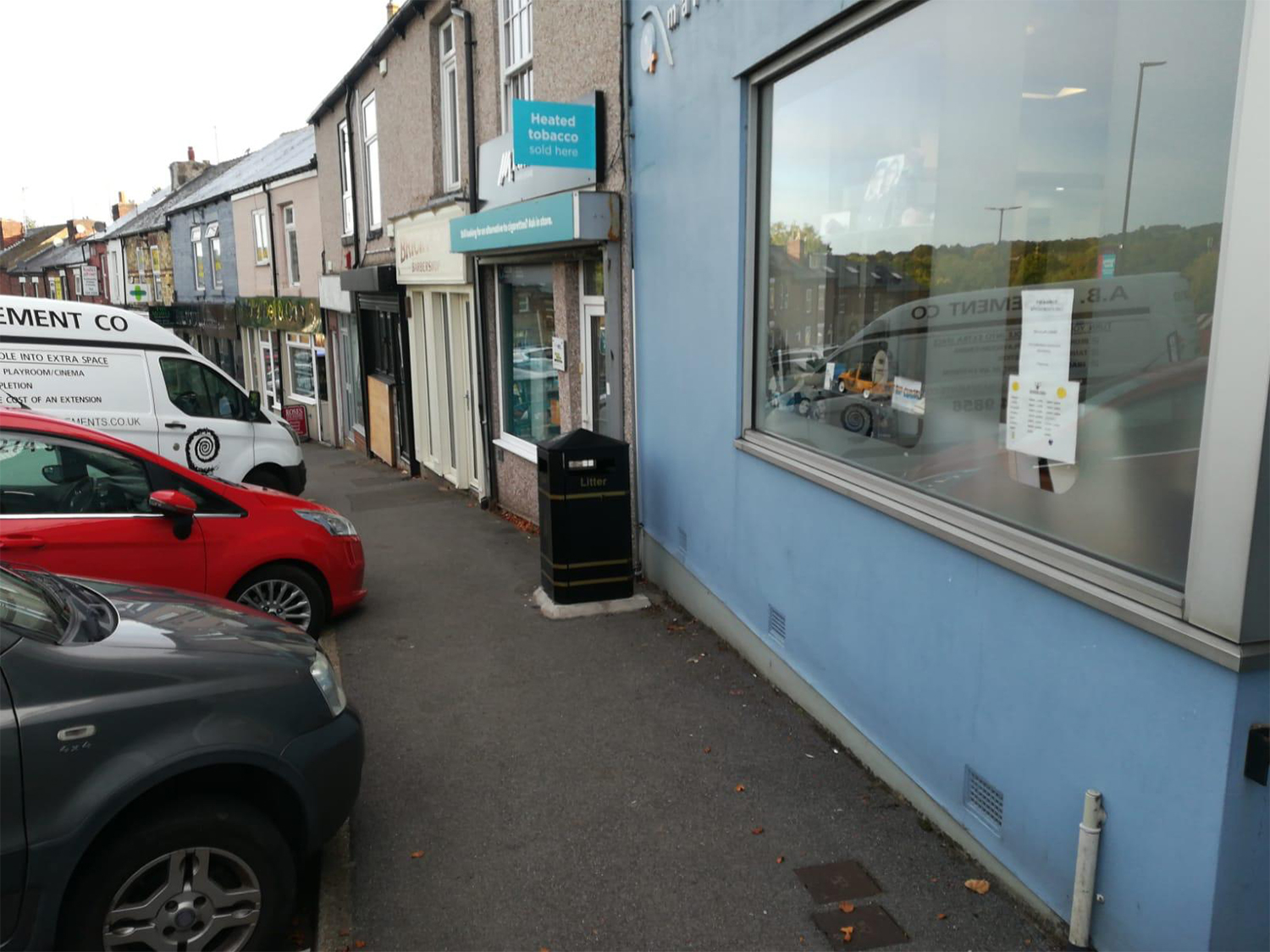 Narrow pavement alongside a row of small shops, including a store with a sign reading “Heated tobacco sold here.” A black litter bin is positioned near the shop entrance. Parked vehicles, including a red car and a white van with signage, line the street on the left. The foreground shows a blue-painted building with a large window displaying a notice, and reflections of cars are visible in the glass.
