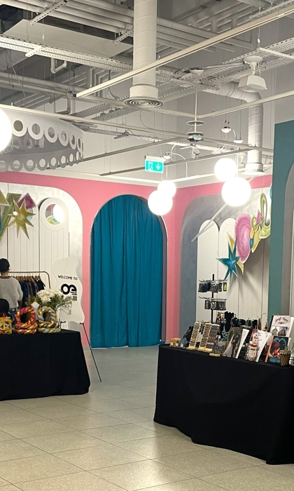 Wide view of a pop-up market interior with tables covered in black cloth displaying handmade crafts, artwork, and clothing. The space features vibrant wall decorations in pink, teal, and yellow with large hanging lights.