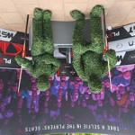 Two Hedge Men street performers dressed as human-shaped living topiary sit at a small table, holding sticks, with event signage and a crowd behind them.