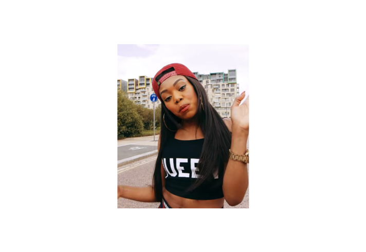 Portrait photo of a woman standing outdoors on a street with modern apartment buildings in the background. She has long dark hair, wears a red baseball cap backwards, large hoop earrings, a black crop top with white lettering, and a gold watch. She tilts her head and raises one hand in a playful, expressive gesture while looking towards the camera.