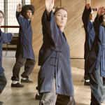 A group of people practise slow, coordinated martial arts movements in a bright wooden hall. Wearing loose navy-blue training uniforms with black trousers and shoes, participants stand in staggered rows with one arm raised and the other extended, holding balanced stances. Large leaded windows and wood-panelled walls form the backdrop, while sunlight falls across the polished wooden floor.