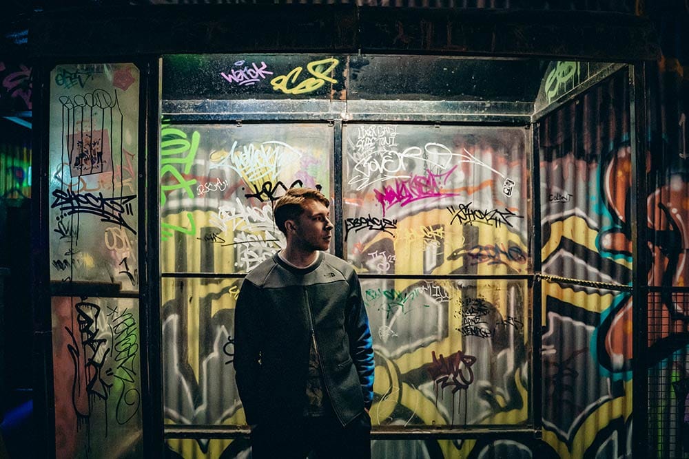 Person standing inside a graffiti-covered glass shelter at night, surrounded by colourful tags and street art illuminated by moody lighting.